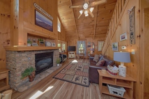 A cozy log cabin interior with a stone fireplace, a comfortable sofa, a television, and a ceiling fan. - Serenity Ridge | Mtn Views, Hot Tub, Near Dollywood, Fireplace + Free Attraction Tickets (Pigeon Forge)