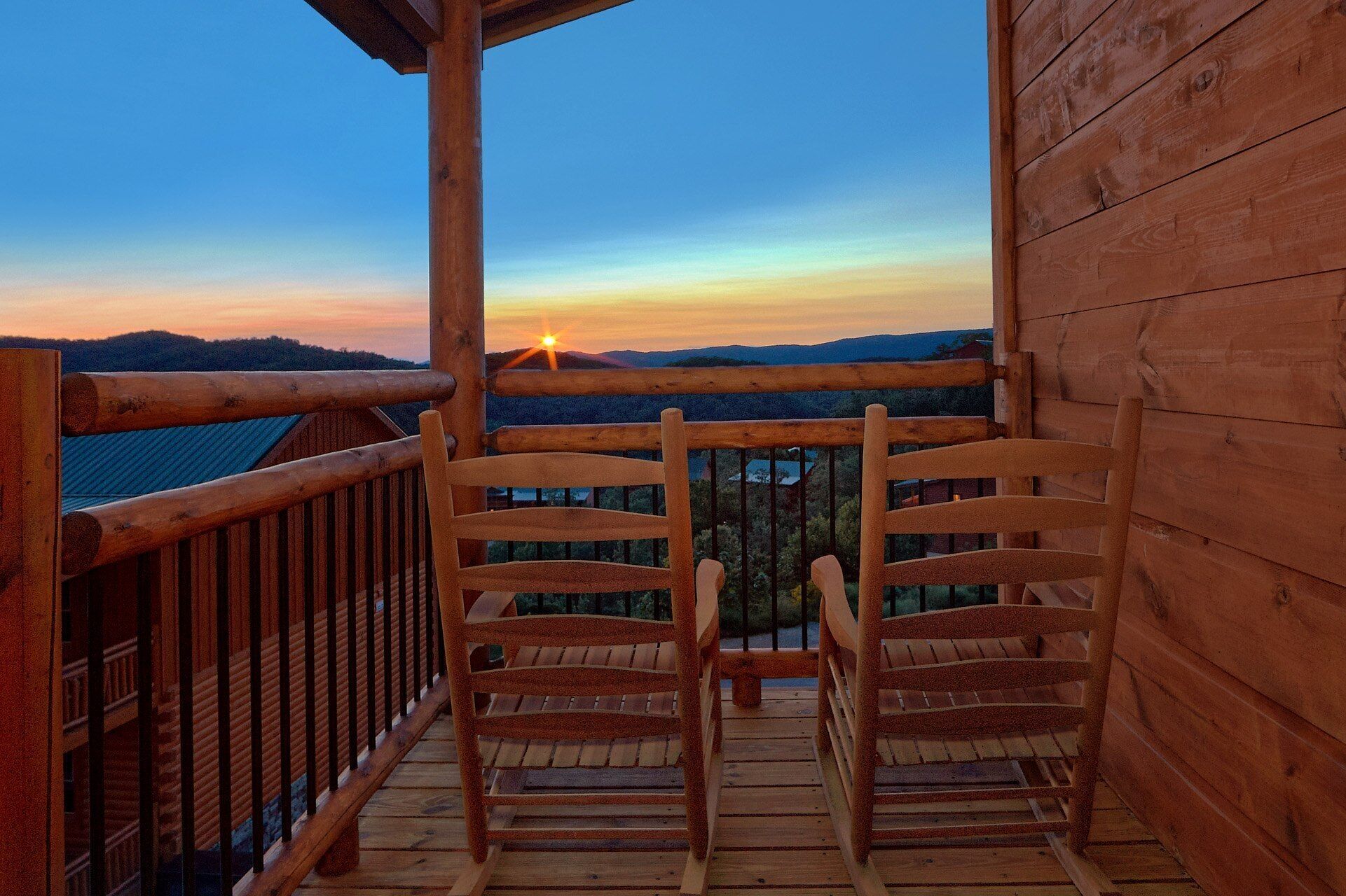 A wooden deck with two wooden chairs facing a scenic view of a sunset over a mountainous landscape.