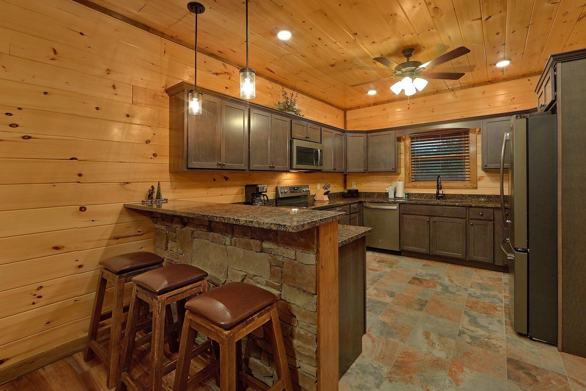 A kitchen with wooden walls, a stone island, bar stools, and a ceiling fan.