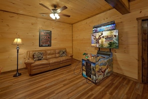 A room with a brown leather sofa, a ceiling fan, a mounted television displaying 'Big Buck Hunter Arcade', and a framed poster on the wall. - Smoky Bear Lodge | Private Pool, Mtn Views, Hot Tub, Game Room, Sleeps 10 + Free Attraction Tickets (Wears Valley)