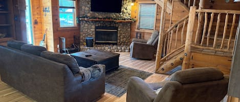 A cozy living room with a stone fireplace, a flat-screen TV, a wooden staircase, and a sofa.
