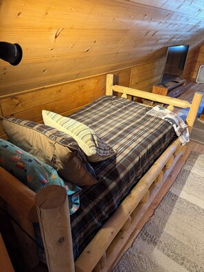 Ein hölzernes Bett mit kariertem Überwurf und Kissen, ein Holz-Kopfstück und ein kleiner Nachttisch mit Lampe.