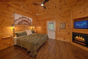 A wooden cabin bedroom with a bed, a television, a fireplace, and a ceiling fan. - Smoky Mountain Lodge | Sleeps 17, Theater, Hot Tub, Game Room, King Suites + Free Attraction Tickets (Pigeon Forge)