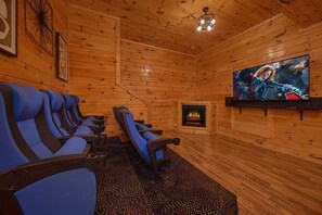 A wooden cabin interior with a fireplace, a television mounted on the wall, and a row of blue chairs. - Smoky Mountain Lodge | Sleeps 17, Theater, Hot Tub, Game Room, King Suites + Free Attraction Tickets (Pigeon Forge)