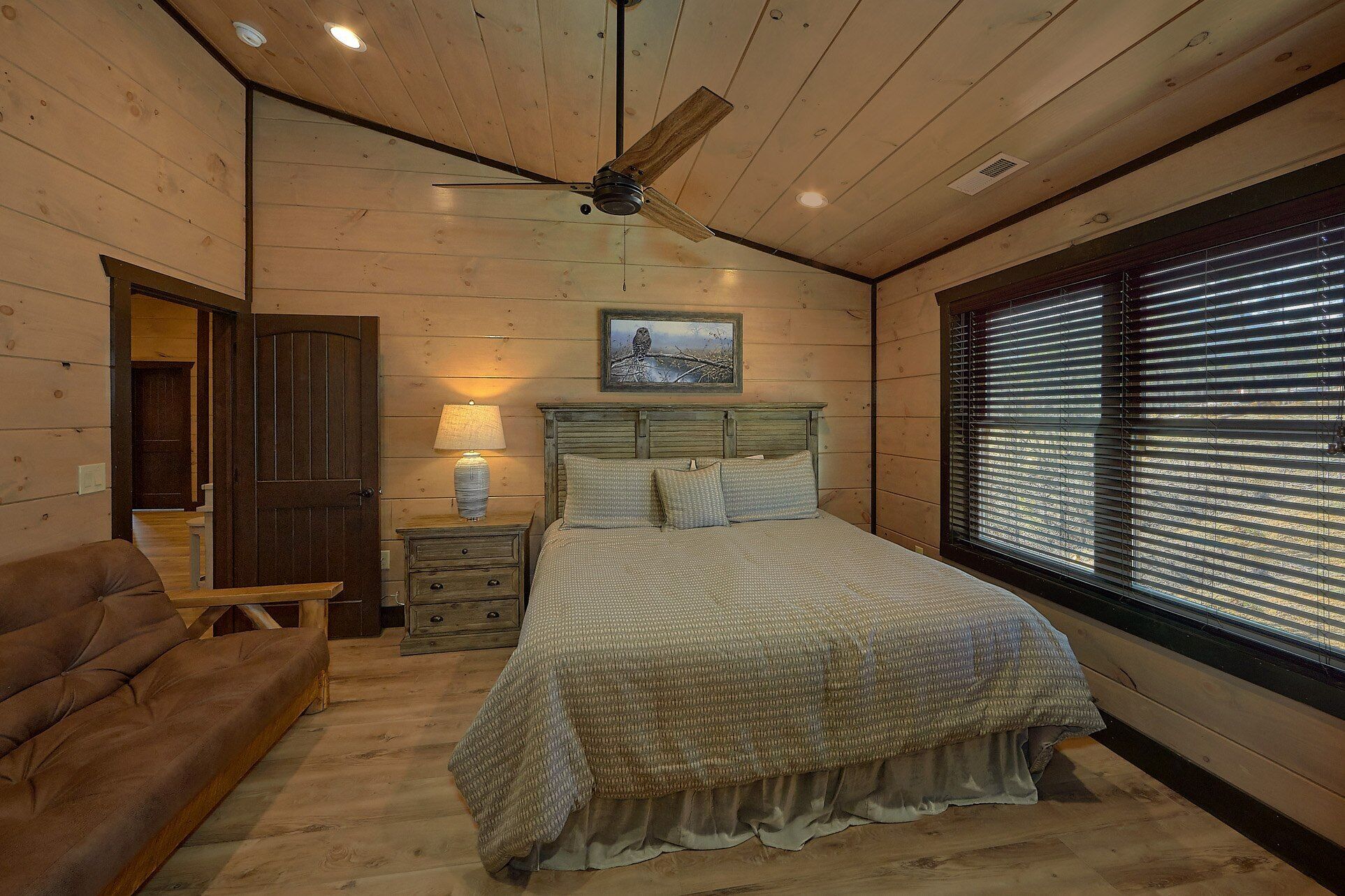 A bedroom with a wooden ceiling, a bed with a headboard, a nightstand with a lamp, a window with blinds, and a door.