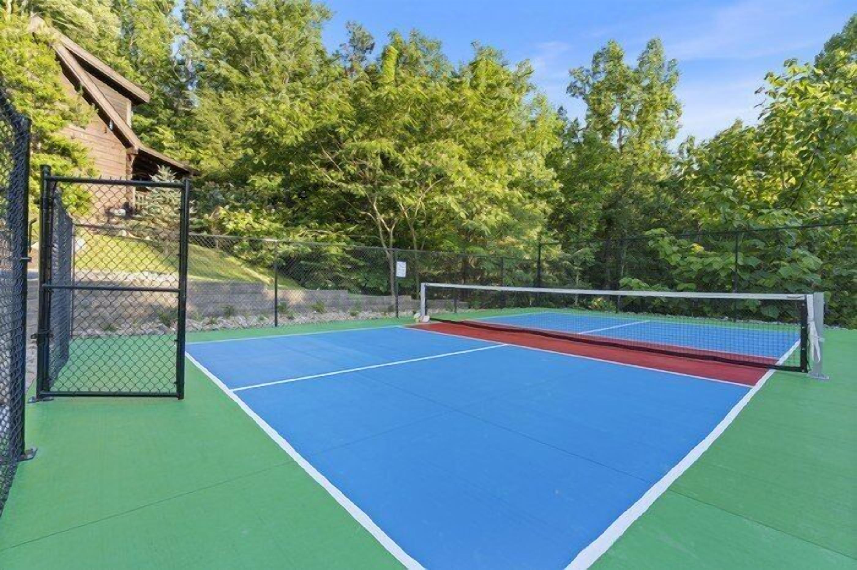 Una cancha de tenis al aire libre con red, rodeada por una cerca y árboles.