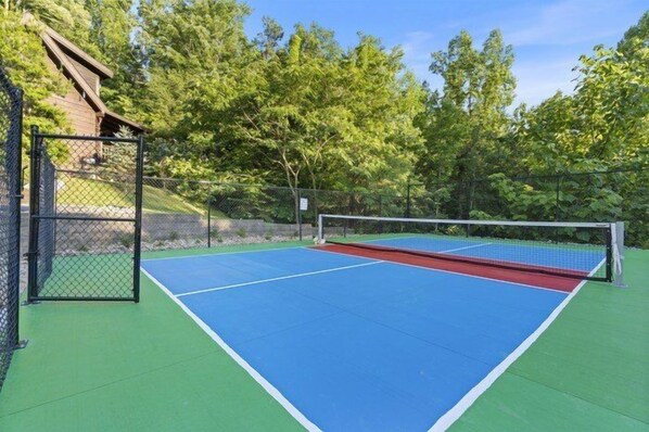 An outdoor tennis court with a net, surrounded by a fence and trees. - Splashin On Smoky Ridge | Private Pool, Theater, Smoky Mtn Views, Hot Tub + Free Attraction Tickets (Wears Valley)