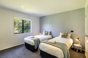 A bedroom with two beds, a window with a view of trees, a bedside lamp, and a small round table. - Riverside Retreat (Taupo)