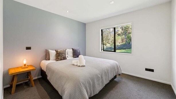 A bedroom with a bed, a bedside table with a lamp, and a window with a view of greenery. - Riverside Retreat - spa pool near the riverside (Taupo)