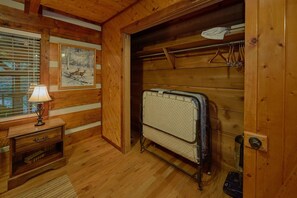 A wooden room with a bed, a bedside table with a lamp, a framed picture on the wall, and a coat rack with hangers. - Two Cubs Den | Arcade, Fireplace + Free Attraction Tickets (Sevierville)