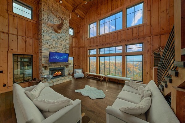A spacious living room with a stone fireplace, a mounted deer head, and a large window offering a view of autumn foliage. - Sunshine Mountain Vista | Private Pool, Mtn Views, Game Room, Sleeps 10 + Free Attraction Tickets (Gatlinburg)