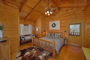 Ein Schlafzimmer in einer Holzhütte mit einem Bett, einer Holzkommode, einem Spiegel, einem Fenster mit Jalousien, einem Deckenventilator und einer Tür mit Fenster.