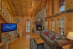 A log cabin interior with a stone fireplace, wooden bookshelves, a television, and a sofa with decorative pillows. - Heavenly-RAE Cabin Rental (Wears Valley)