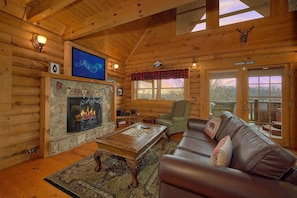 A log cabin interior with a stone fireplace, a television mounted on the wall, a leather sofa, and a wooden coffee table. - A Romantic Hilltop | Hot Tub | Pool Table (Sevierville)