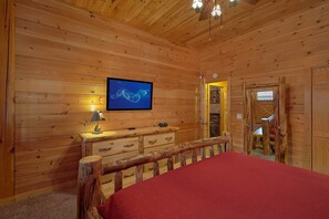 A wooden cabin room with a bed, a television on the wall, a dresser, and a door leading to another room. - Mountain Retreat Cabin Rental (Wears Valley)