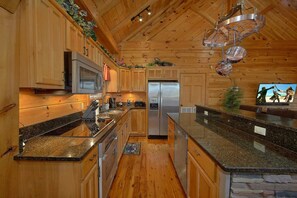 A kitchen with wooden cabinets, stainless steel appliances, granite countertops, and a stone accent wall. - Mountain Retreat | Theater, Mtn Views, Hot Tub, Fireplace + Free Attraction Tickets (Wears Valley)