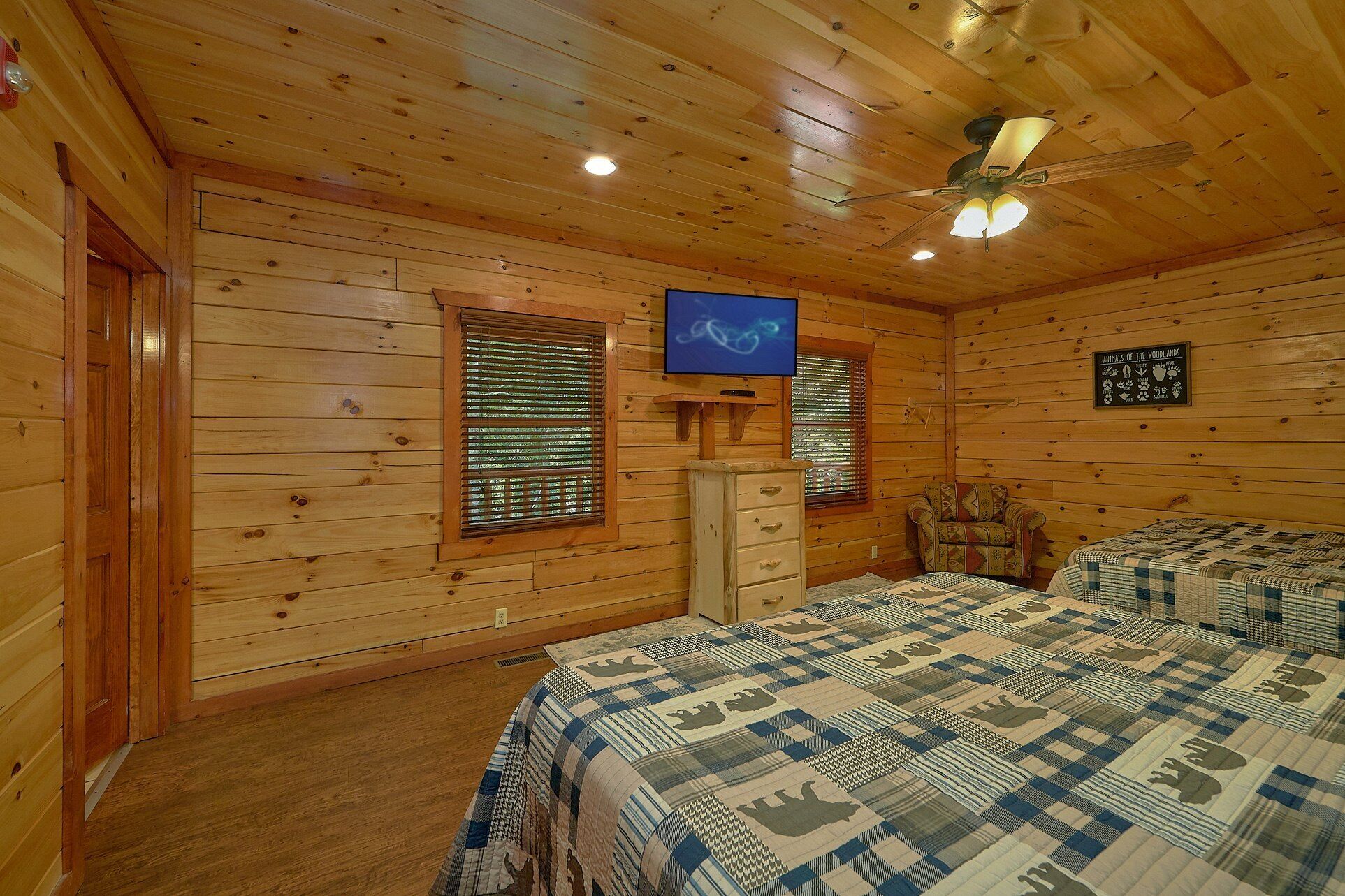 Une chambre de style cabane en bois, équipée de deux lits, d’un ventilateur de plafond, d’une télévision et d’une étagère fixée au mur.