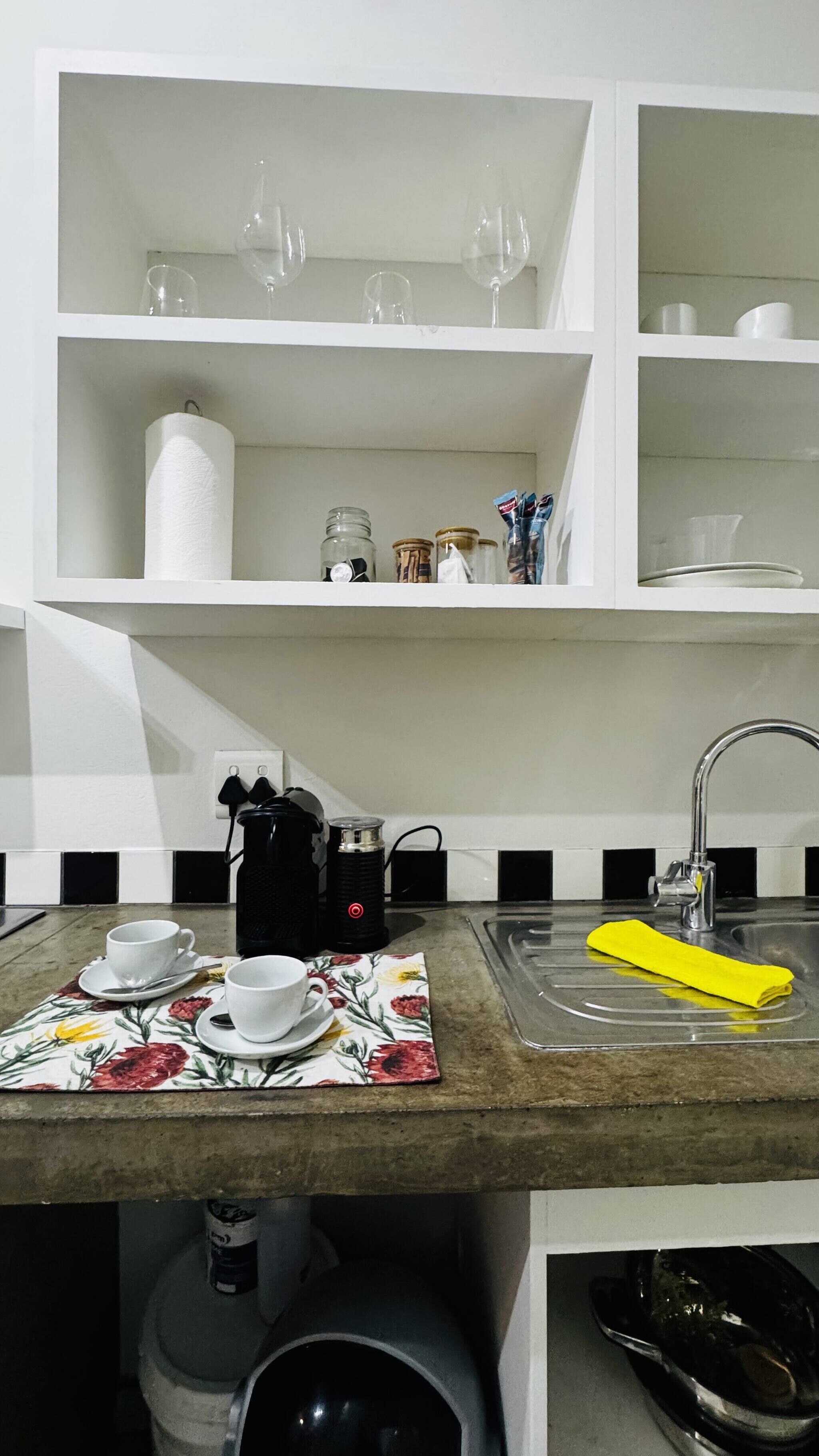 Cocina con armarios blancos, un lavamanos y una encimera con cafetera, tazas y una toalla amarilla.