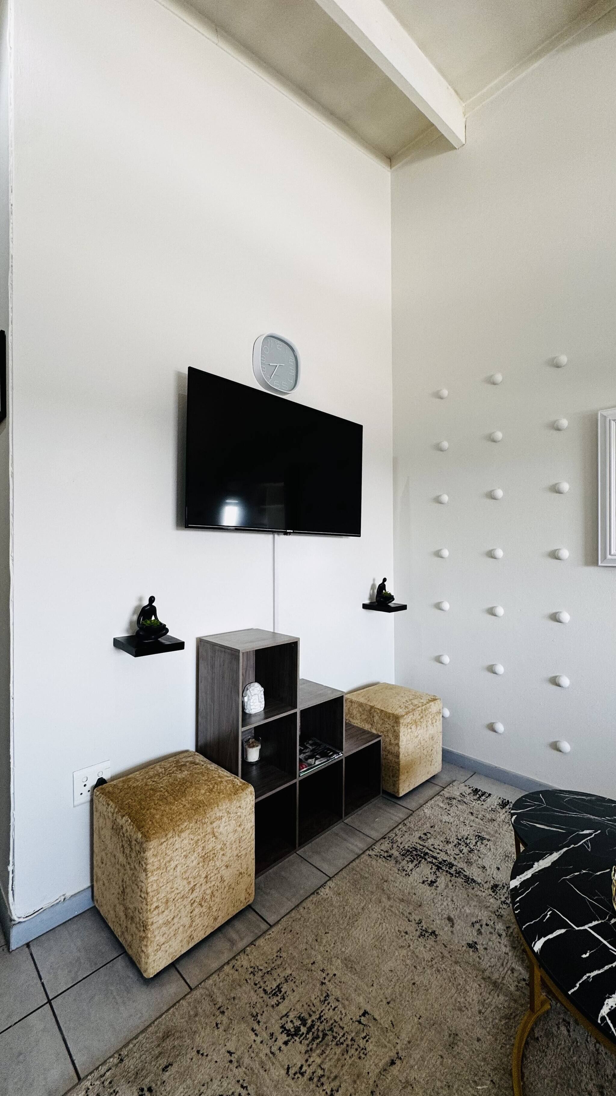 A modern living room with a flat-screen TV mounted on the wall, a small shelf with decorative items, a wooden cabinet, and two square ottomans.