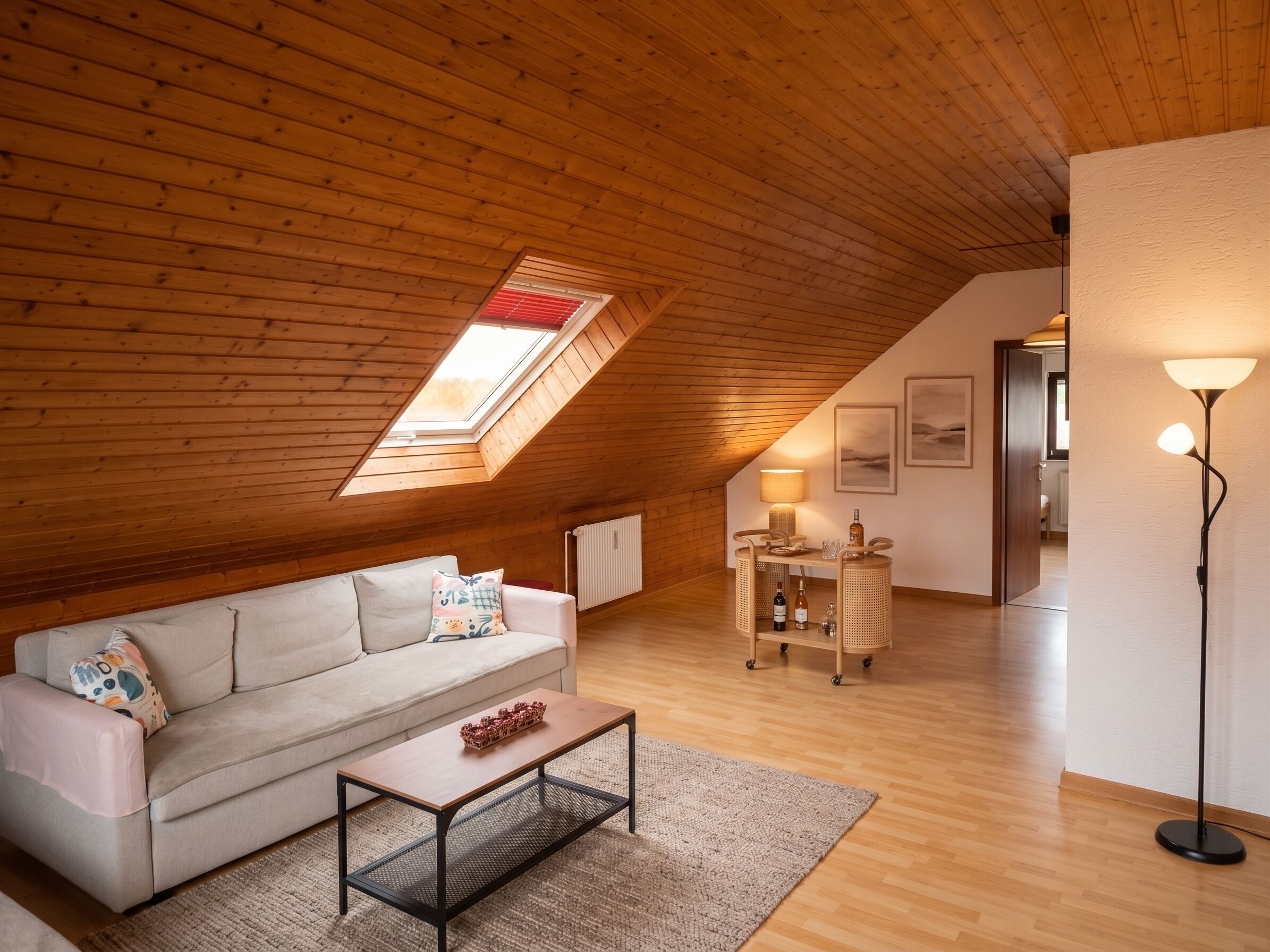 Une chambre mansardée chaleureuse, avec un plafond en pente, une lucarne, un canapé, une table basse, un bureau et une lampe sur pied.