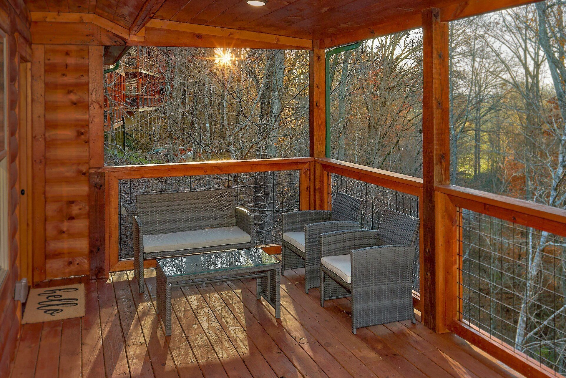 A wooden deck with wicker furniture and a glass table, surrounded by a metal fence, overlooking a forested area.