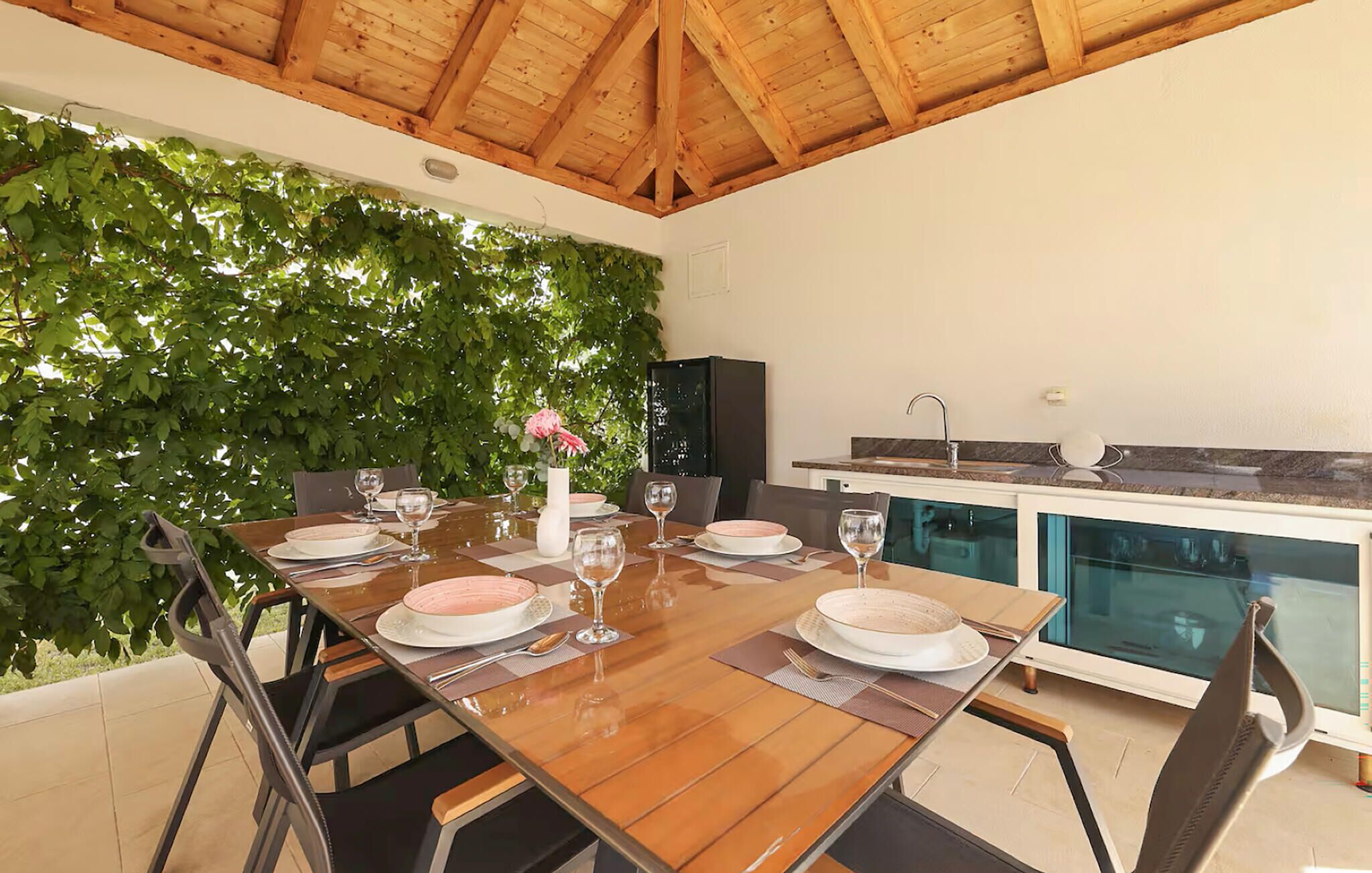 Un espace extérieur aménagé pour les repas, avec une table en bois pour quatre personnes, entourée de chaises, et en arrière-plan, on aperçoit un coin cuisine et une piscine.