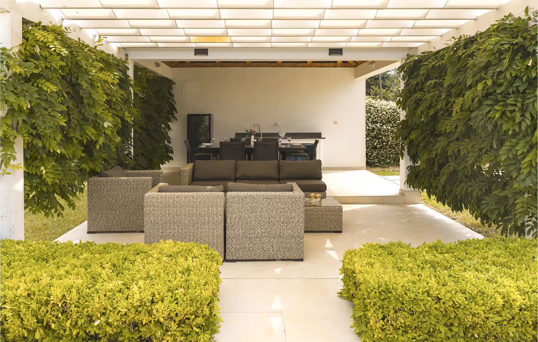 Un espace extérieur aménagé avec un canapé et des fauteuils, entouré de verdure et surmonté d’une pergola.