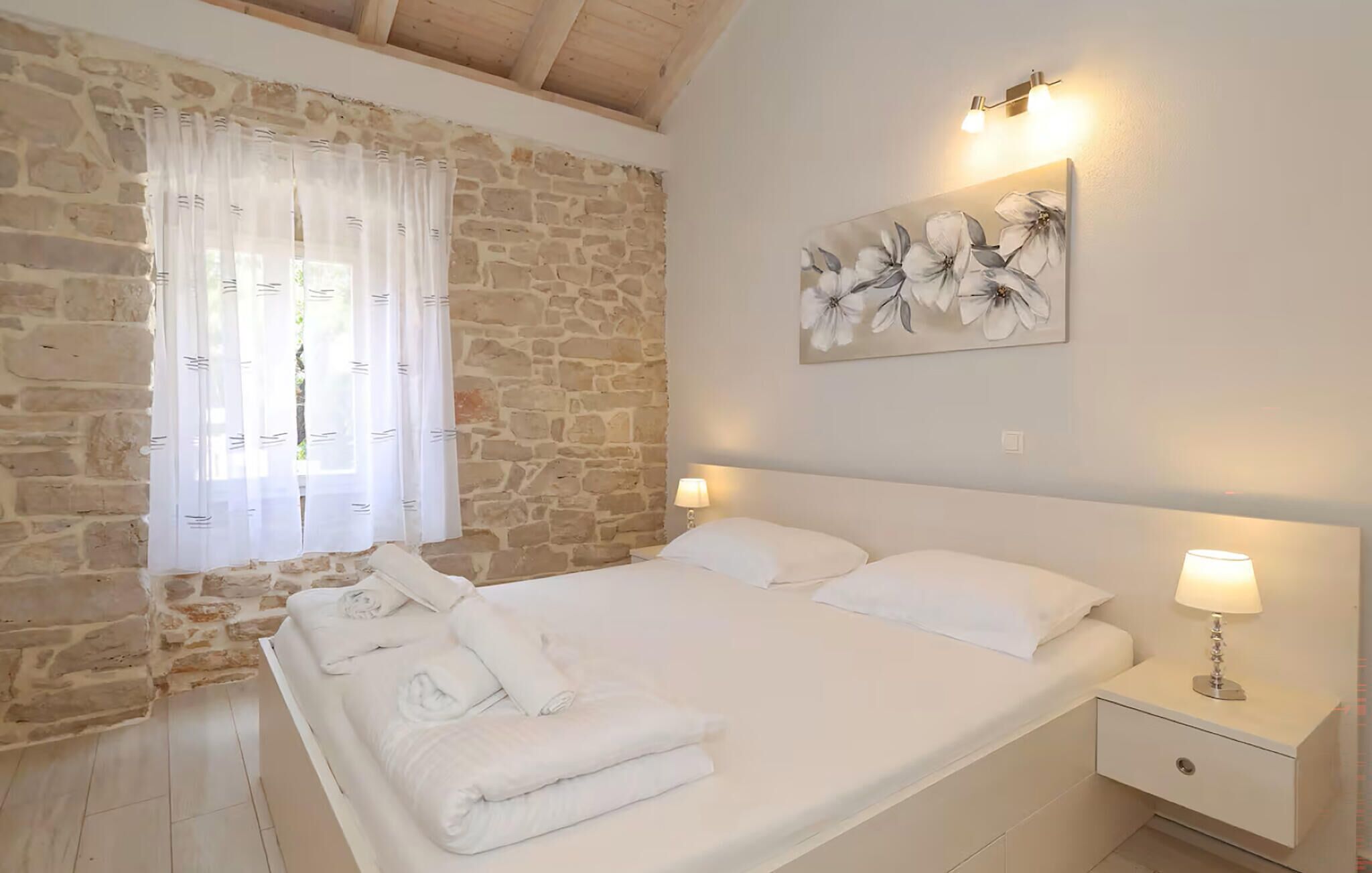 Un dormitorio con pared de piedra, cama con ropa blanca, dos lámparas de noche y un cuadro floral en la pared.