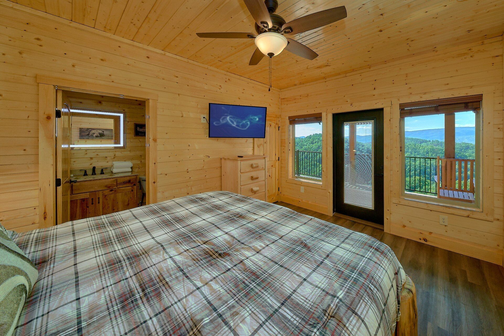 A wooden cabin bedroom with a bed, a ceiling fan, a window with a view, and a bathroom with a mirror and sink.
