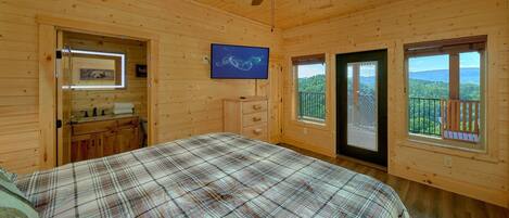Un dormitorio tipo cabaña de madera con cama, ventilador de techo, ventana con vistas y baño con espejo y lavamanos.
