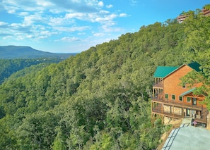 A wooden cabin with a green roof, surrounded by dense forest and hills.