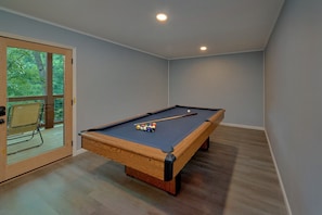 A pool table with balls and a cue stick, a chair, and a door leading to a balcony with a view of trees. - Douglas Dream | Smoky Mtn Views, Hot Tub, Lakefront, Game Room, Fireplace + Free Attraction Tickets (Sevierville)