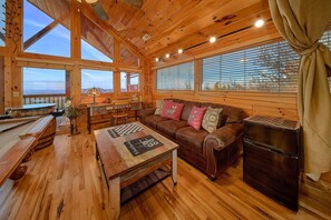 A cozy cabin interior with a wooden floor, a sofa with decorative pillows, a table with a chessboard, and large windows with blinds. - Chocolate Moose | Mountain Views | Hot Tub | Pool Table (Sevierville)