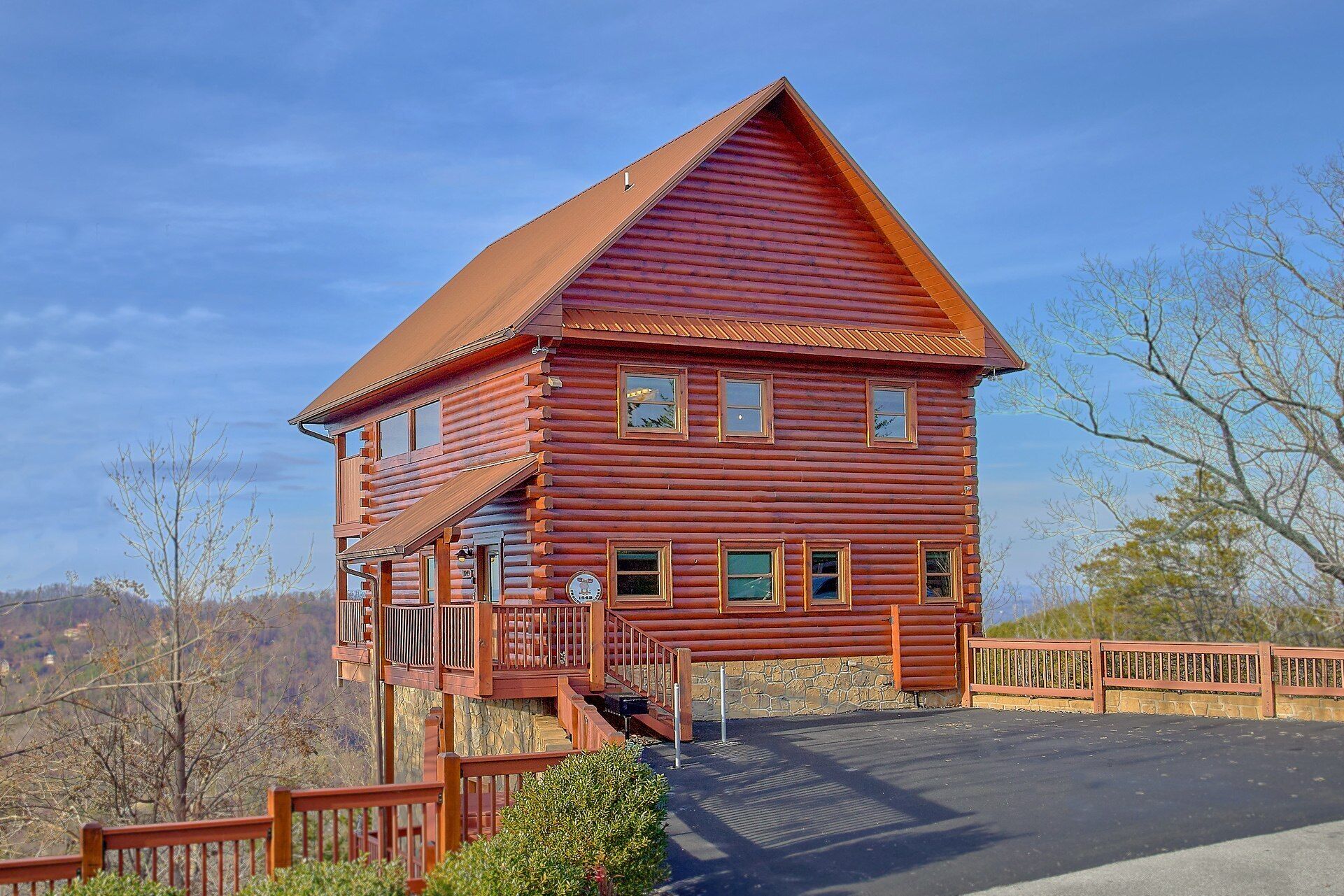 A two-story log cabin with a balcony, a stone foundation, and a fenced driveway.