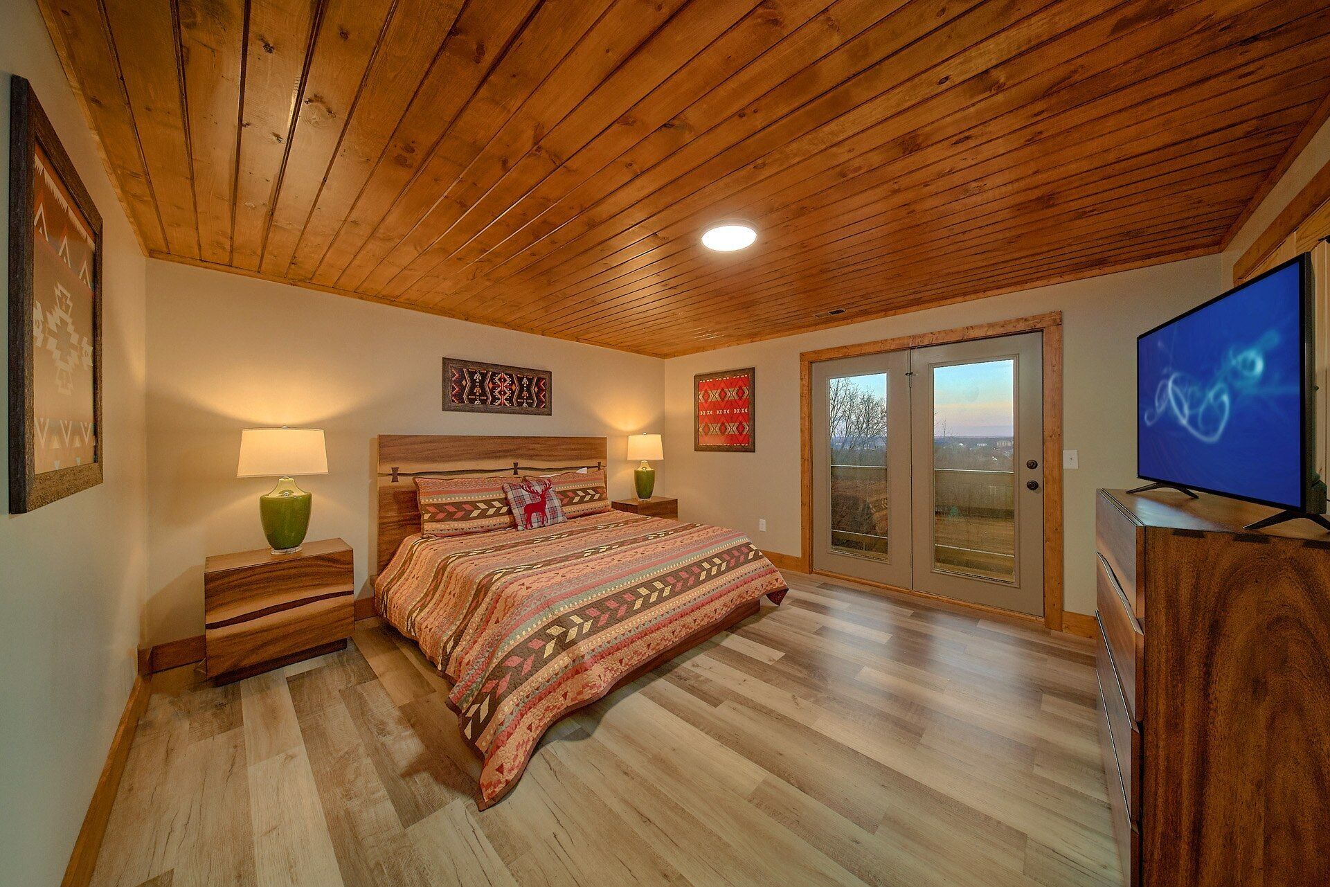 A bedroom with a wooden ceiling, a large bed with patterned bedding, two bedside lamps, a television, and a sliding glass door leading to a balcony.