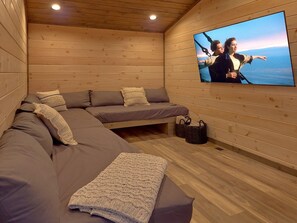 Ein gemütliches Zimmer mit einem großen Flachbildfernseher an der Wand, einer Holzbalkendecke und einem Ecksofa mit Kissen.