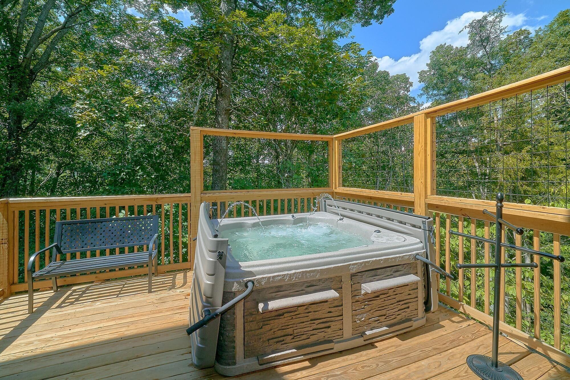 A hot tub on a wooden deck with a bench, surrounded by a wooden railing and trees.