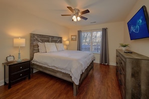 A bedroom with a wooden bed, bedside lamps, a ceiling fan, a TV, and a window with curtains. - Bear Necessities Vacation Home (Sevierville)