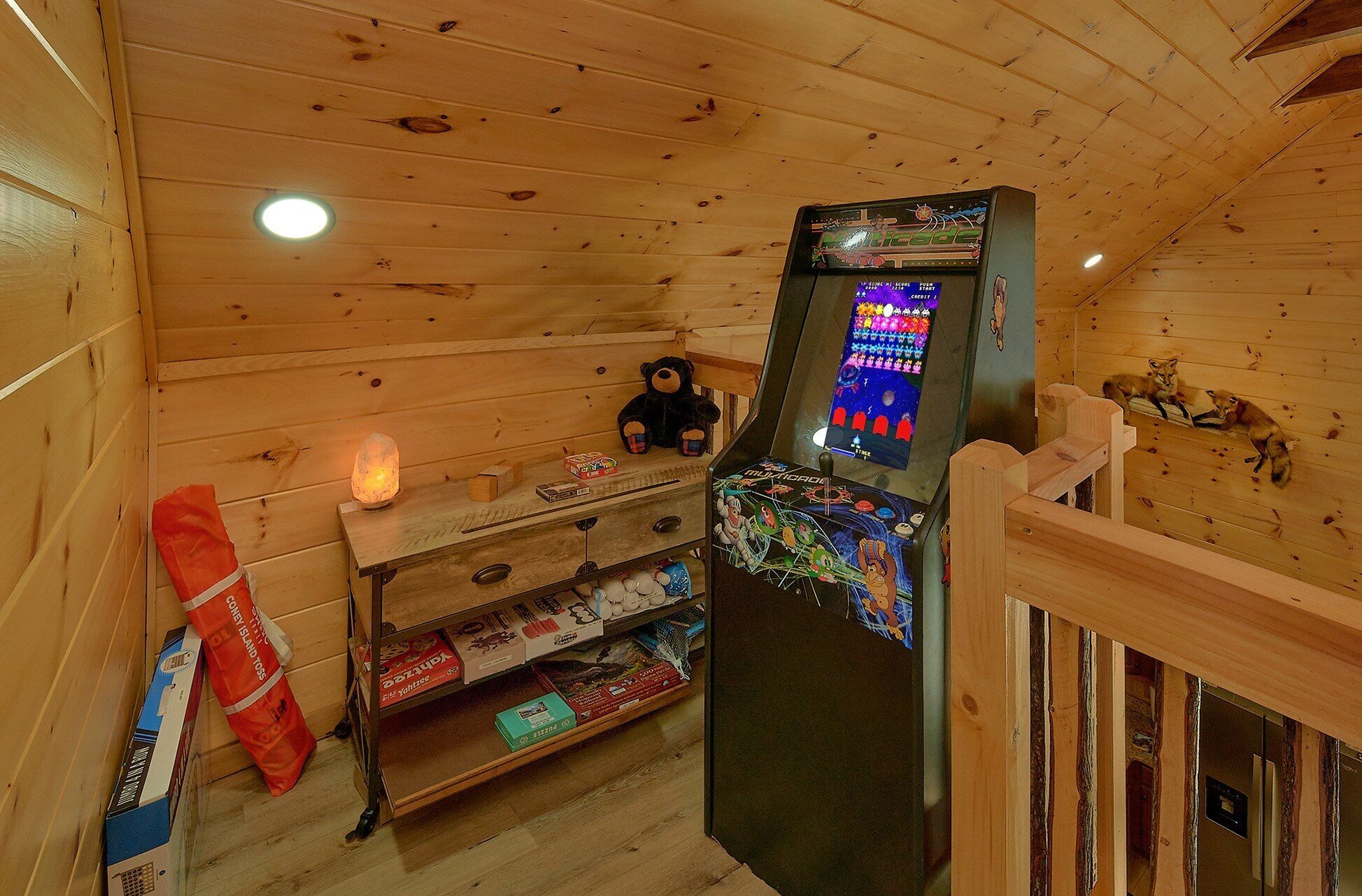 Interior de una cabaña de madera con una consola de videojuegos, una estantería con juegos de mesa y una zona de estar acogedora.