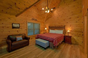 Een knus kamer in een blokhut met houten plafond, een bed met roodruitig sprei, een bruine leren bank, een raam met jaloezieën en een plafondventilator.