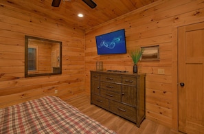 A wooden cabin interior with a bed, dresser, mirror, and a television mounted on the wall. - Smoky Vista Lodge | Theater, Mtn Views, Hot Tub, Game Room, Sleeps 10 + Free Attraction Tickets (Gatlinburg)