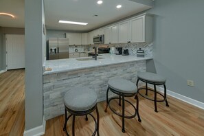 A kitchen with a granite countertop, stainless steel appliances, and a stone backsplash. There are three bar stools with cushioned seats. - Mountain View 2504 | Mtn Views, Pigeon Forge, Resort Pool, Fireplace + Free Attraction Tickets (Pigeon Forge)
