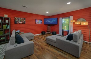 Ein Wohnzimmer mit roten Wänden, einem Flachbildfernseher, zwei grauen Sofas, einem Holzstuhl und einem Bücherregal.