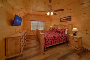 A wooden cabin bedroom with a bed, nightstands, a dresser, a ceiling fan, a television, and a window with blinds. - Makin' Memories | Hot Tub, Game Room, Fire Pit, Fireplace + Free Attraction Tickets (Sevierville)