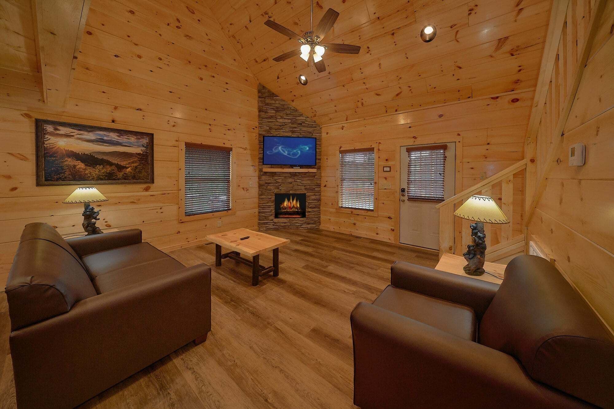 A log cabin interior with a fireplace, two sofas, a TV, and a ceiling fan.