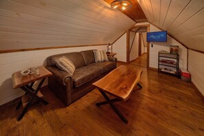A cozy attic room with a brown leather sofa, a wooden bench, and a small table with a lamp. A television is mounted on the wall, and there are board games on a shelf. - Jewell's Place | Fire Pit, Pet Friendly, Fireplace + Free Attraction Tickets (Sevierville)