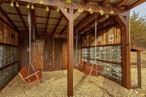 A rustic wooden structure with two swings suspended from the ceiling, surrounded by wooden walls and a gravel floor. - Jewell's Place | Fire Pit, Pet Friendly, Fireplace + Free Attraction Tickets (Sevierville)