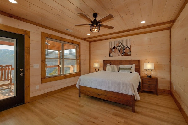 Un dormitorio con techo de madera, una cama, dos mesitas de noche con lámparas, una ventana con persianas y un ventilador de techo.