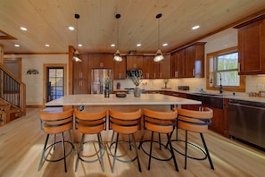 Una cocina con isla central, taburetes de bar, armarios de madera, electrodomésticos de acero inoxidable e iluminación colgante.