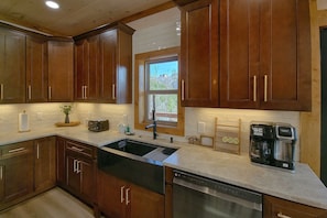 Una cocina con armarios de madera, lavavajillas de acero inoxidable, cafetera y ventana con vistas a árboles.